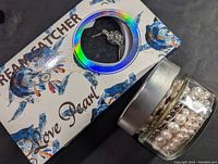 Glass jar filled with loose drilled pearls next to a blue decorative gift box with a clam pendant visible through a circular window.