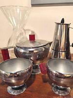 Full set photo showing chrome 3-piece tea service with red bakelite handles alongside glass coffee carafe and stovetop espresso moka pot on wooden surface.