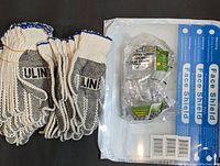 Overview photo showing several pairs of U-Line knit gloves, packaged clear plastic safety goggles, and multiple packaged full face plastic shields laid out on a black surface.
