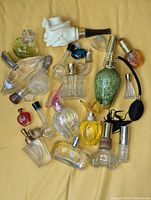 Top-down photo showing 19 perfume and cologne bottles in assorted sizes, shapes, and colors including a white sculptured face bottle, green bottle with atomizer bulb, and various heart-shaped and geometric clear glass bottles.