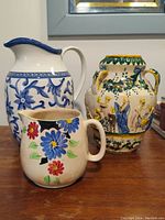 Lot display showing the three pieces: two pitchers (one large with blue floral design, one smaller with multi-color flowers) and a larger hand-painted Italian ceramic vessel with figures and two handles.