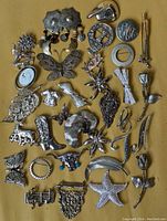 Top down photo of 34 silver tone brooches in various shapes and sizes arranged on a beige fabric background.