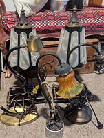 Photo showing all assorted vintage lamps together on carpeted floor against furniture backdrop. Contains two tall black lamps with white plastic shades and finials, two brass gooseneck lamps, and one small yellow-orange glass flower lamp with metal base.