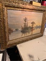 Frontal view of vintage oil painting showing landscape with water, trees, and islands, framed in an ornate gold frame.