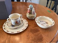 Photo showing two-handled Bunnykins soup mug, two plates with bunny border patterns, and a Bunnykins soup bowl on wooden table.