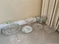 Full lot showing all glass cake stands and glass bowl arranged on carpet near curtain and wall corner.