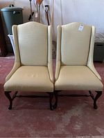 Two matching beige fabric wingback chairs side by side, showing full front view of upholstery and wooden legs.