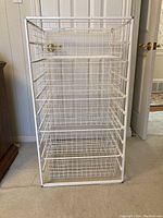 Front view of white metal wire closet organizer with five empty adjustable baskets, placed on carpeted floor near a door.
