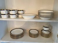Six teacups, oval serving bowl, stack of rimmed plates displayed on upper shelf of cabinet.