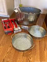 Photo showing two new Weber BBQ tool sets each containing a spatula and tongs on red packaging, next to three galvanized aluminum pans including one large bucket and two smaller handled pans, placed on wooden floor.