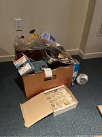 Cardboard box filled with assorted electrical items and containers with light bulbs, including original packaging of some bulbs.