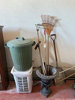 Wide view showing the full lot contents including green plastic trash can with lid on the white plastic laundry basket, decorative garden planter urn containing various garden tools including rakes, shovel and a hoe.