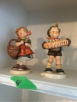 Pair of Goebel Hummel figurines on a shelf with green tape indicating 5 inches in height.