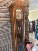 Front and side view of wooden grandfather clock placed against textured stone wall near the floor. Shows tall case, carved floral details above the clock face, and visible brass weights and pendulum inside glass door.