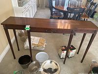Front view of a long rectangular wood hall/console table with three tapered legs visible and carved decorative details along the edges under the top surface. Scratches and water damage on the top are evident.
