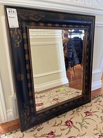 Framed mirror shown standing on carpeted floor leaning against a wall, displaying the ornate black and gold frame with floral and striped detailing.