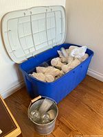 Large blue plastic storage bin open showing wrapped plastic stemware inside, with a galvanized metal bucket in front containing clear glass cups and pitchers.