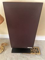 Front view of one speaker covered with brown fabric mesh, sitting on black rectangular stand on floor.