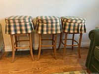 Front view of three wooden kitchen stools with plaid cushioned tops and rattan seats.