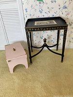 Front view of black wooden table with gold floral designs and lithograph inset on top next to pink wooden bench showing size relation and overall look.