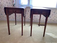 Pair of vintage wood drop leaf tables with drawers shown standing closed side-by-side on carpeted floor in front of window.