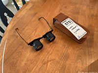 Magnifying glasses positioned next to the original brown leather case with label indicating 'watch maker's glasses'. Shows glasses frame, lenses, and case details.