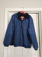 Front view of the blue LL Bean fleece lined full zip jacket hung against a white door. Shows ribbed cuffs, ribbed waistband, and collar with fleece lining visible inside.