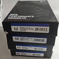 Photo of the boxes showing size labels and style names for four Nike swimwear items; three visible labels: Poly Blend Color Fade Jammer size 22, Core Solids Brief size 28, and other swimwear sizes.