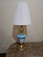 Full view of the vintage table lamp showing white pleated shade, blue jasperware body with decoration, and shiny brass base on tiled surface.