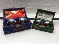 Two pairs of polished metal massage balls housed separately in two fabric boxes, one blue and one green. The green box includes an information card about the product.