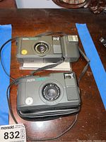 Two Agfa Compact cameras shown from top angle on wooden surface. Cameras are black with various lens markings and a camera strap is visible. One camera sits atop a black pouch.