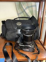 Canon PowerShot S3 IS camera with attached lens and strap placed in front of black camera bag on wooden shelf.