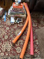 Stack of board games and playing cards with foam pool noodles on a carpeted floor, including Monopoly and Parcheesi, along with other boxed games.