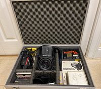 Aluminum hard case open showing camera body, lens, extension tube box, meter, filters, cable, empty box and other accessories in foam insert