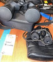 Two Nikon binoculars on a wooden surface, one larger with Nikon case, the smaller with lens covers and enclosed in a carrying pouch.