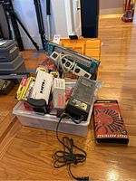 Various tools including battery testers, battery charger, surge suppressor, tape measures, and other small tools organized in a plastic bin on a wooden floor.
