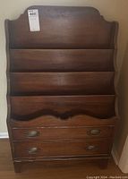 Front view of antique wooden magazine rack showing three tiered shelves and two drawers with brass handles.