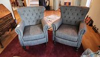 Front view showing pair of upholstered plaid wingback armchairs with button tufted backs and rolled arms on a red rug.