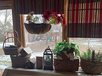 Photo showing a collection of baskets, a hanging basket planter with artificial flowers, a black metal lantern with glass panels, and a square woven basket with artificial greenery on a table by a window.