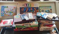 Photo showing an assortment of vintage and modern board games and puzzles stacked together on a surface. Games include The Game of Life, Sorry!, Yahtzee, Stratego, and Candy Land among others, plus puzzle sets and card decks.