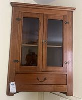 Front view of the wooden corner wall cabinet showing two glass doors and drawer below.
