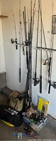 Group of fishing rods leaning against wall with reels attached plus rod case and tackle box on floor
