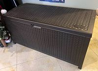 Closed Suncast outdoor storage bin showing woven rattan-style plastic surface and lockable handle.