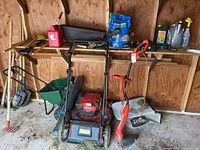 Wide shot showing lawn mower, trimmer, seed spreader, wheelbarrow, hand tools, gas can, and lawn care materials in shed.