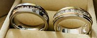 Close-up of two men's rings, showing one two-tone ring with dark and clear stones and one single-tone ring with clear stones on a beige jewelry holder.