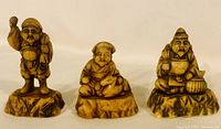 Set of three Asian peasant figurines shown together on a white background, standing and seated poses, carved detail and textured bases visible.