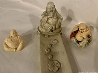 Set of 3 ivory color Buddha figurines including a large seated Buddha on a base and two smaller figures, one painted multicolor, on a plain surface.