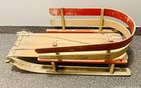 Side view of the vintage child's sleigh showing the red and cream curved wooden rails, flat wooden slatted seat, and attached pulling rope.