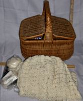 Front view of the wicker basket closed with handle and two flap lids, white wool sweater and knitting supplies in plastic bag in front.