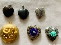 Image shows six heart-shaped lockets and pendants, including two smooth stone pendants (black onyx and tiger eye), multiple silver-tone filigree lockets with floral accents, and a gold-tone oval floral-engraved locket.